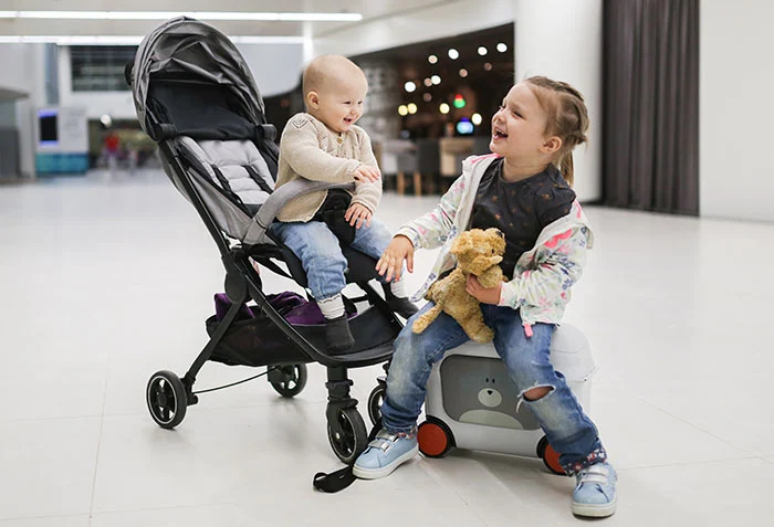Bebis sitter i en liten resevagn på flygplats och skrattar med sin storasyster bredvid