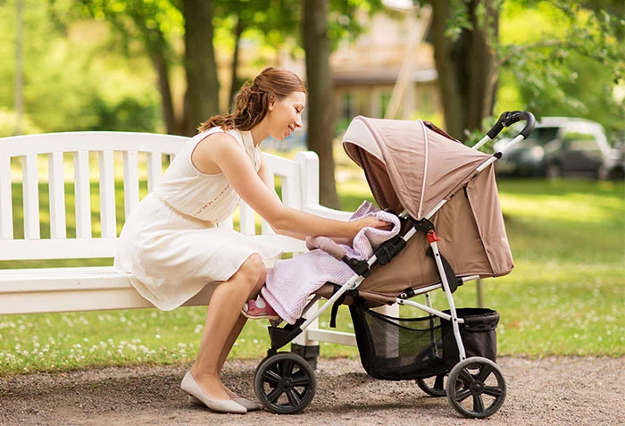 Mamma sitter på parkbänk med barn liggandes i en resevagn bredvid